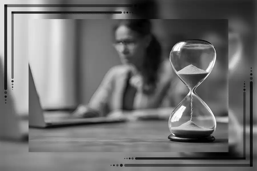 Es ist eine Sanduhr zu sehen, die abläuft. Im Hintergrund sitzt eine Frau, die leicht verschwommen ist. Das Bild ist schwarz-weiß